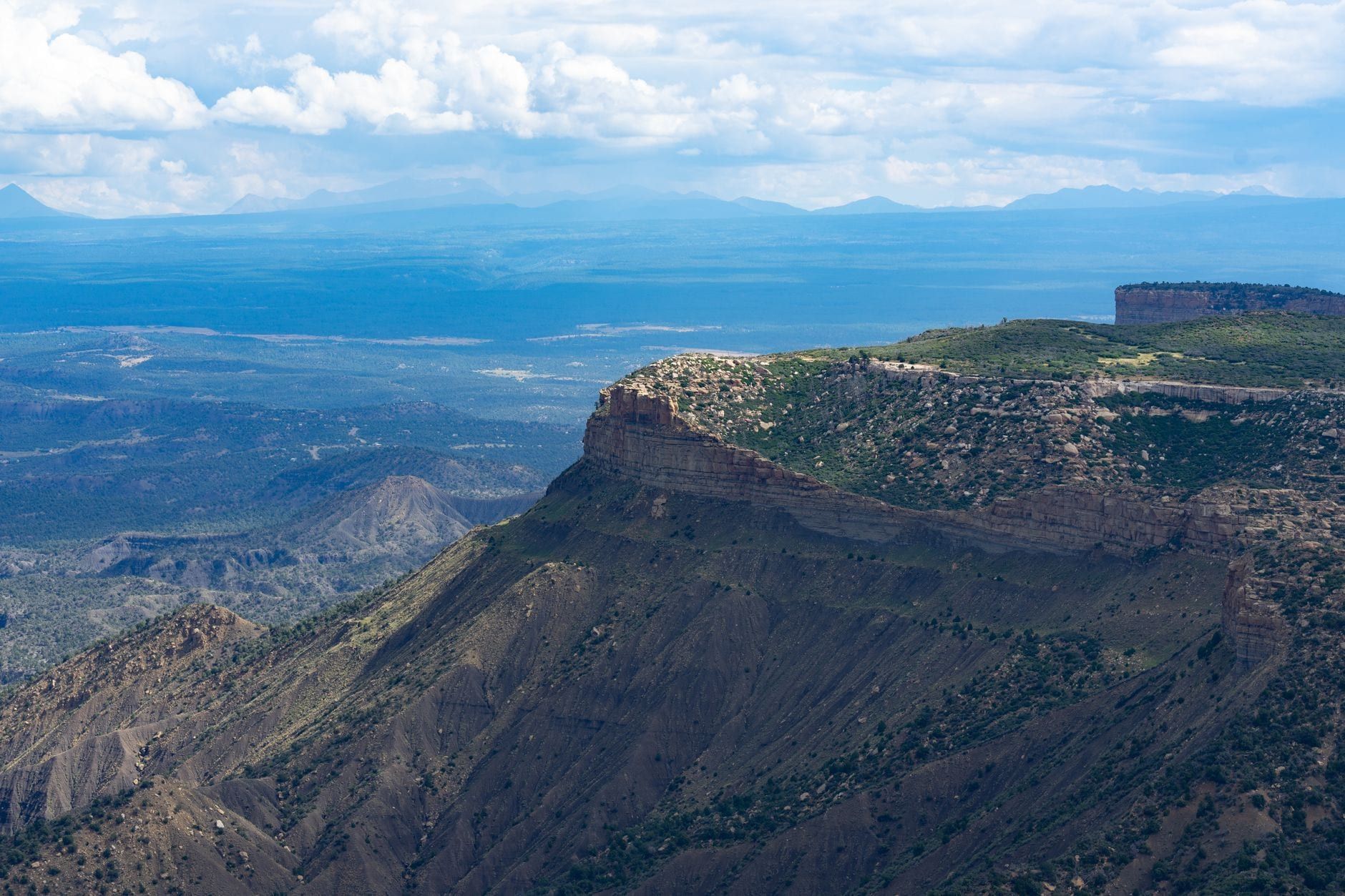 Vista panorámica elevada de los escarpados acantilados y la meseta boscosa del Parque Nacional Mesa Verde extendiéndose hacia el horizonte bajo un cielo parcialmente nublado.